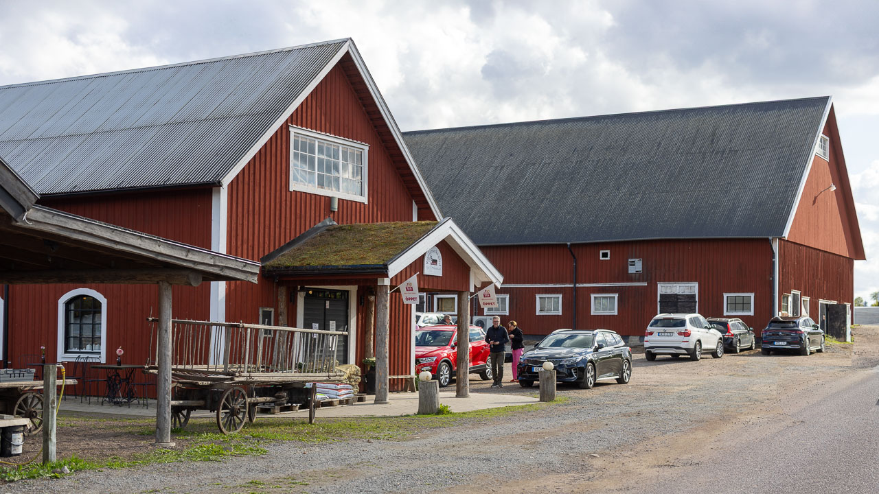 Röd gårdsbyggnad med parkering och bilar utanför under molnig himmel.